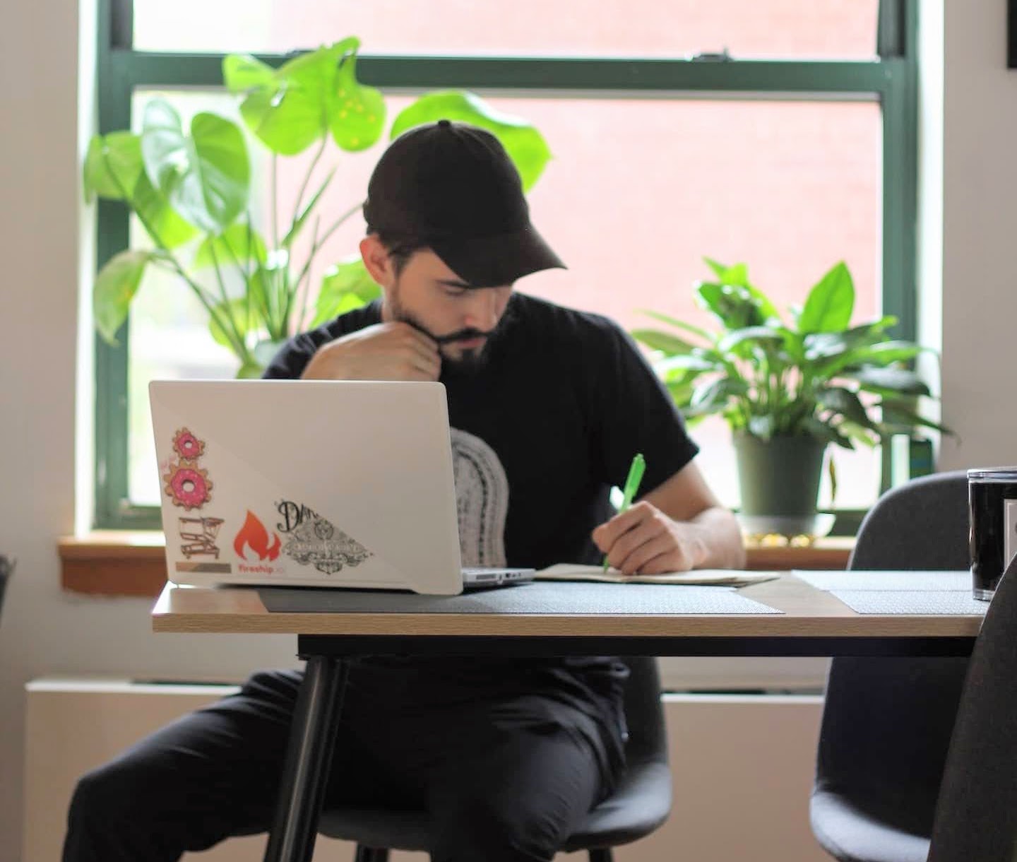 Frank sitting at his dining table, seated in front of a laptop and writing in a notebook. His Monsterra plant and peace lily sit on a window sill behind him.